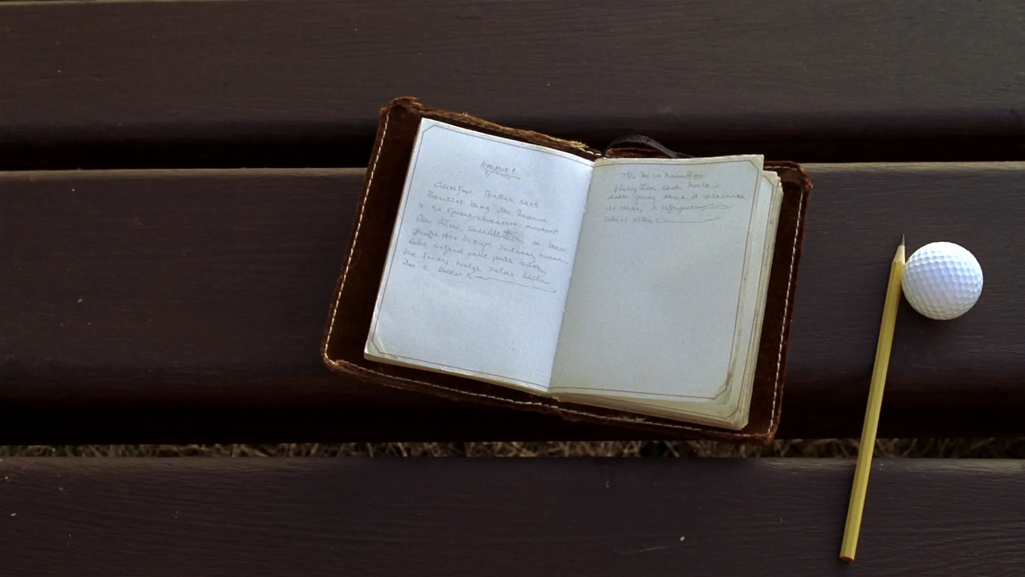 A small leather pocket journal open on a wooden bench beside a golf course with a pencil and a golf ball